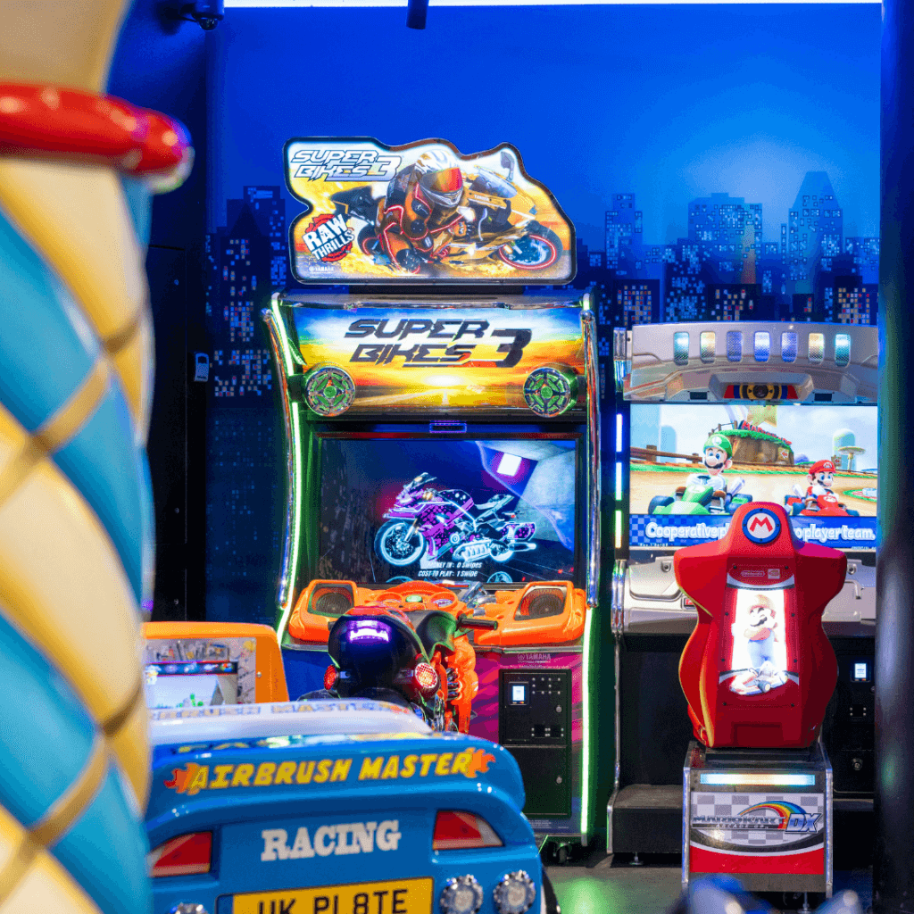 Spielautomaten wie „Super Bikes 3“ und ein Mario-Kart-Rennspiel mit Bildschirmen unter blauem Neonlicht.