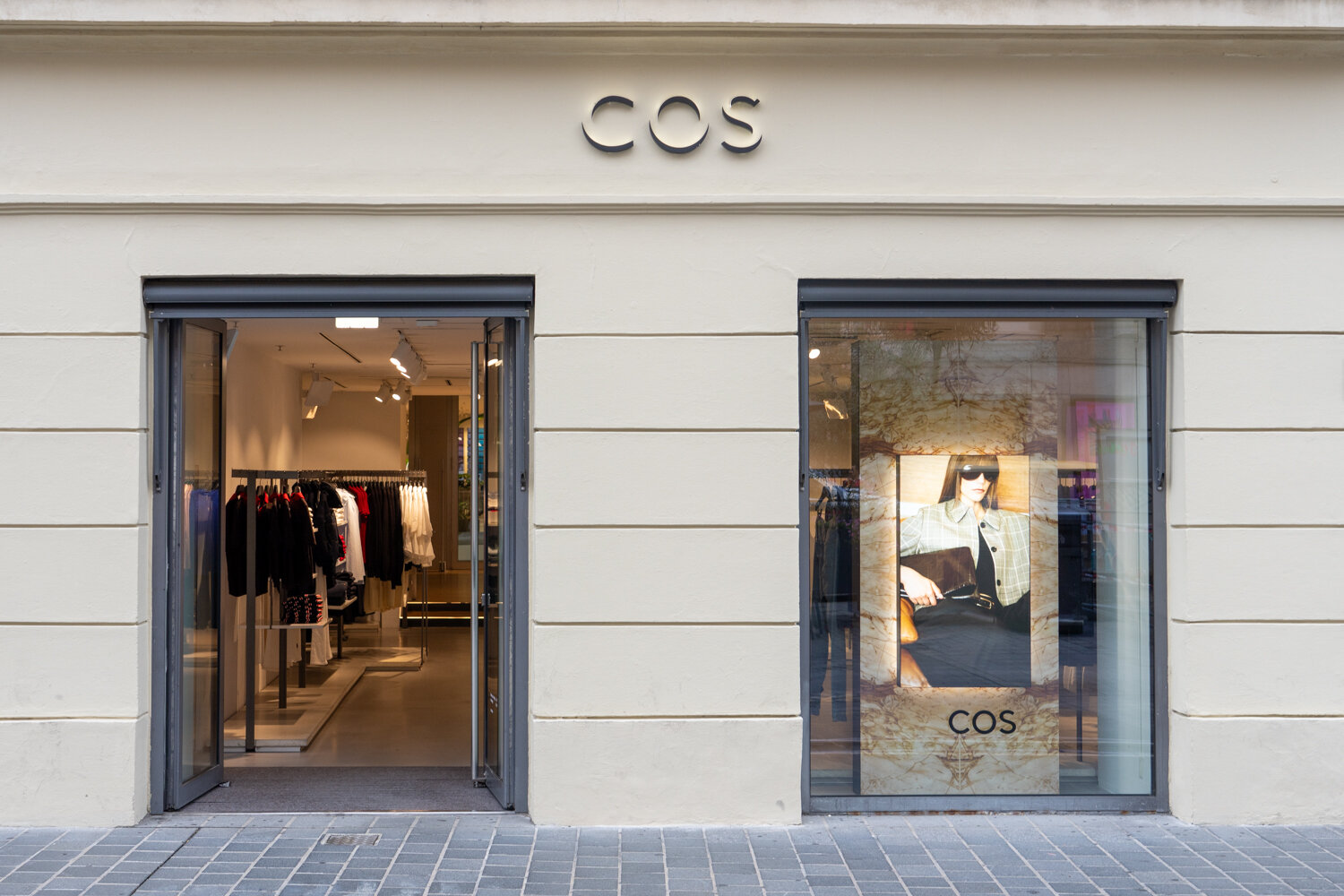 Außenansicht und Eingang des COS Bekleidungsgeschäfts mit hellem Schriftzug und Schaufenster.
