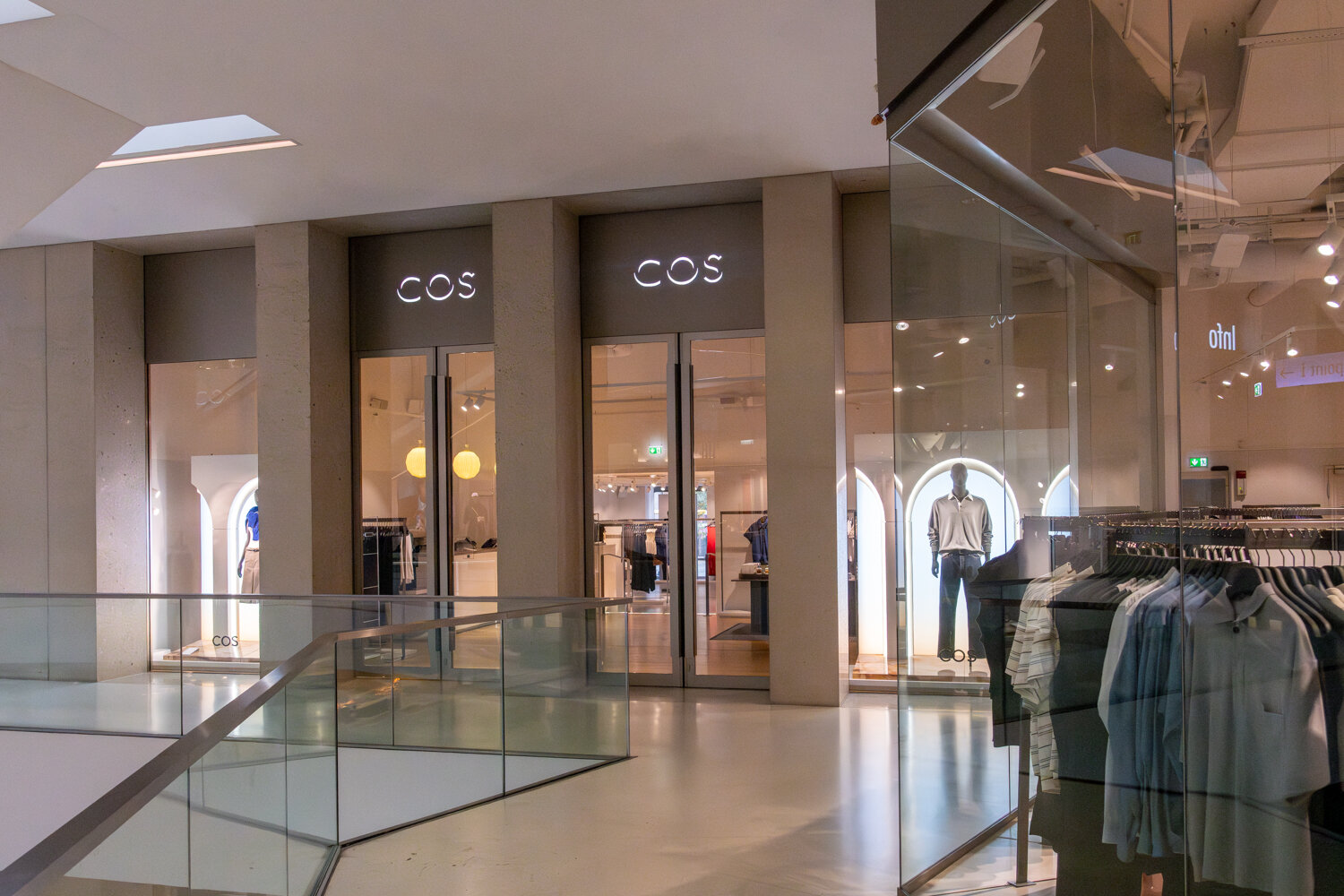 Außenansicht der COS-Filiale im Einkaufszentrum mit Glaseingang, beleuchtetem Markenlogo und Schaufensterpuppen.