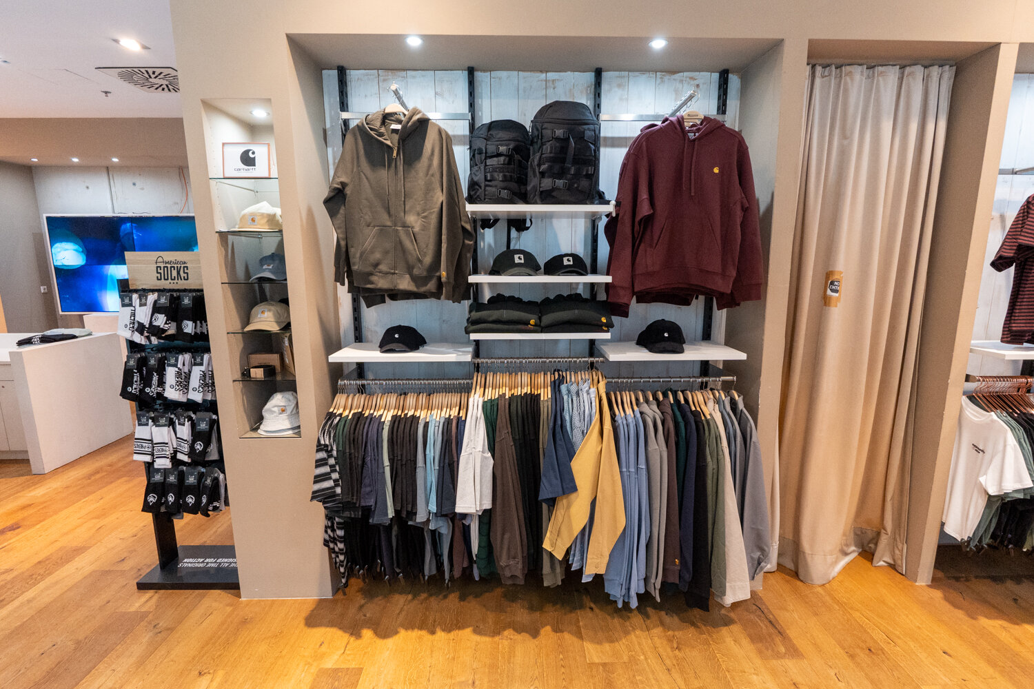 Marken-Wandregal mit aufgehängten Hoodies T-Shirts und Kappen sowie Socken-Ständer und Vorhang zur Umkleidekabine bei Cuprum 29.