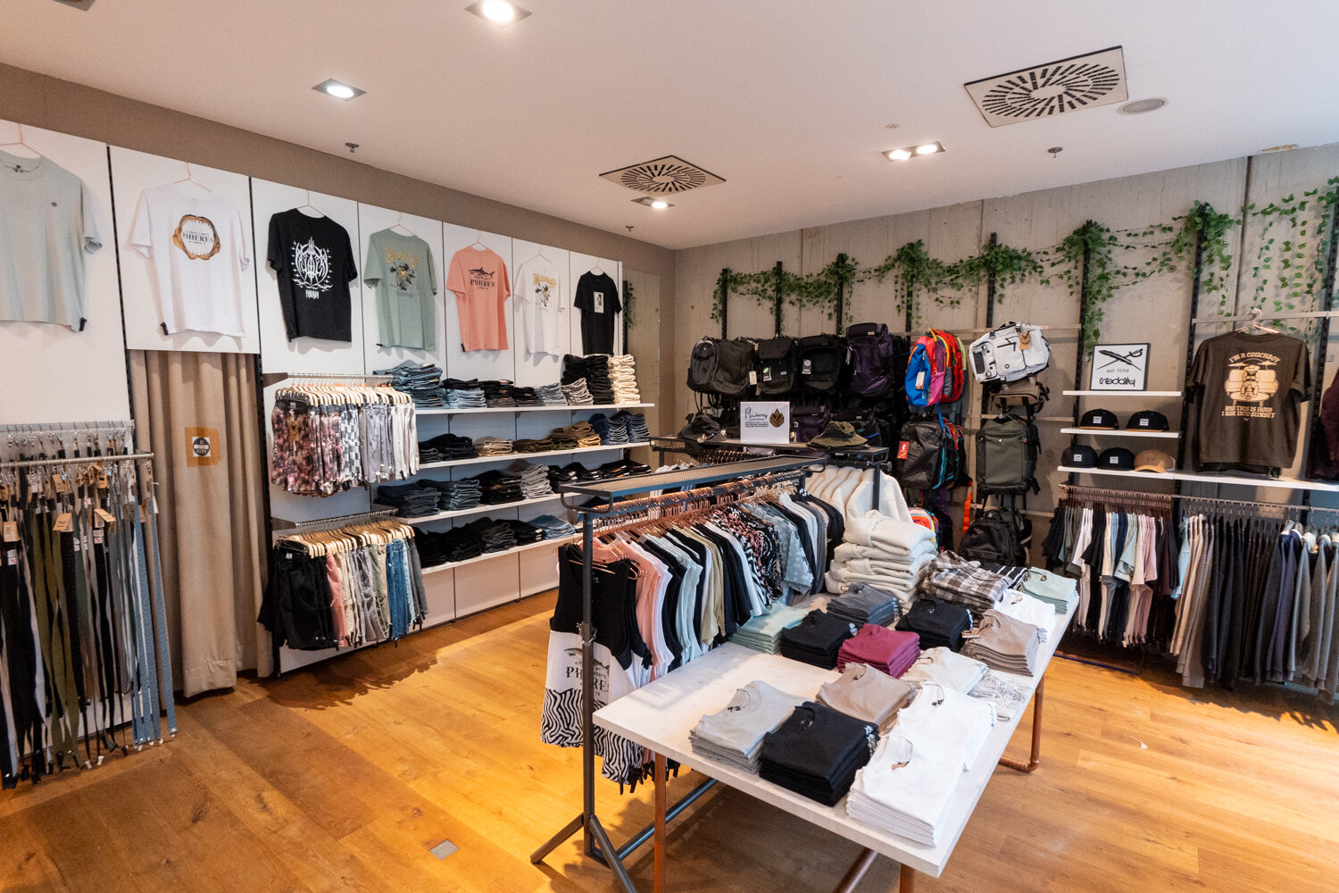 Verkaufsraum mit Kleiderständern voller T-Shirts Hosen und Pullover sowie Regalen mit Rucksäcken und Mützen auf Holzboden im Cuprum29.