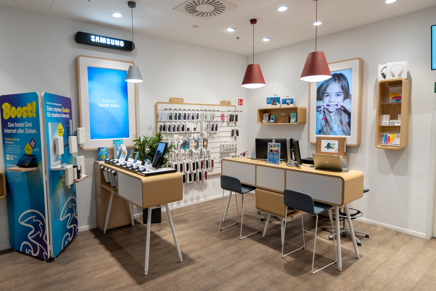 Verkaufsbereich der Drei Filiale mit Ausstellern für Smartphones, Beratungstresen mit Laptops, Hängelampen Zubehörwand und Werbedisplays an der Wand.