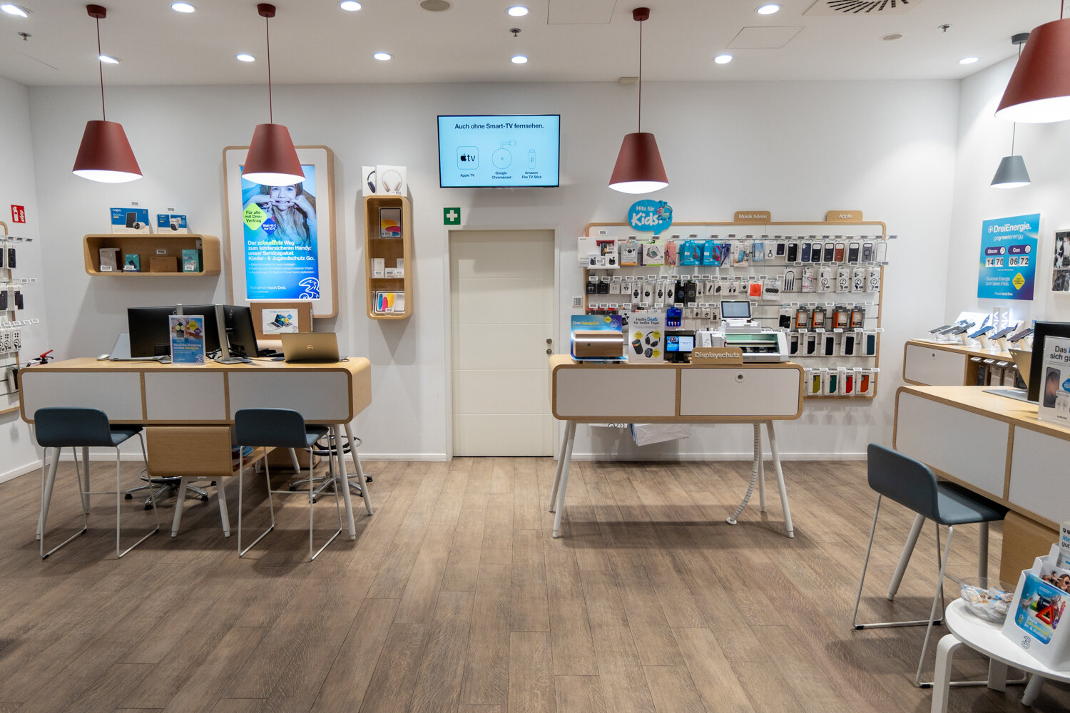 Innenaufnahme des Drei Shops mit hellen Beratungstischen.blauen Stühlen. Zubehörwand für Handyhüllen und roten Hängelampen im Verkaufsraum.