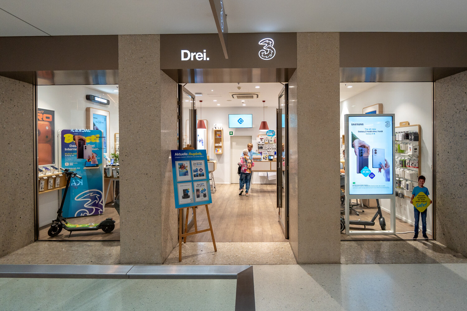 Frontansicht der Drei Filiale mit beleuchtetem Logo über dem Eingang, einem Aufsteller für aktuelle Angebote, Werbedisplays und Blick in den Verkaufsraum.