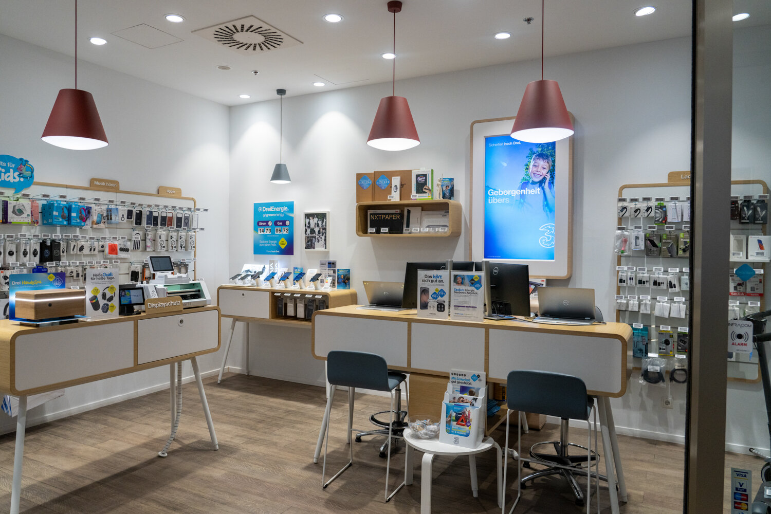 Verkaufs- und Beratungsbereich in einem Drei Shop mit Holzmöbeln, Hängeleuchten, Ausstellungsgeräten und Zubehörwänden.