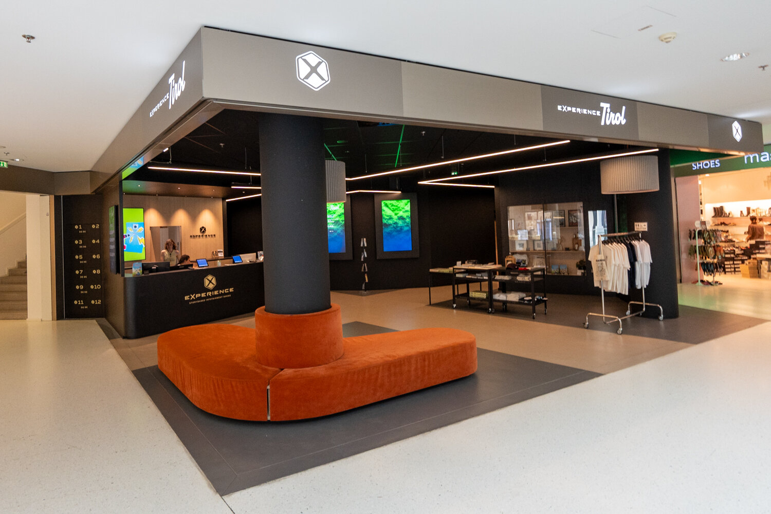 Frontaler Blick auf den Eingang von Experience Tirol mit leuchtendem Schriftzug, orangefarbenem Polstersitz, Empfangstresen, T-Shirt-Ständer und Werbe-Screens.