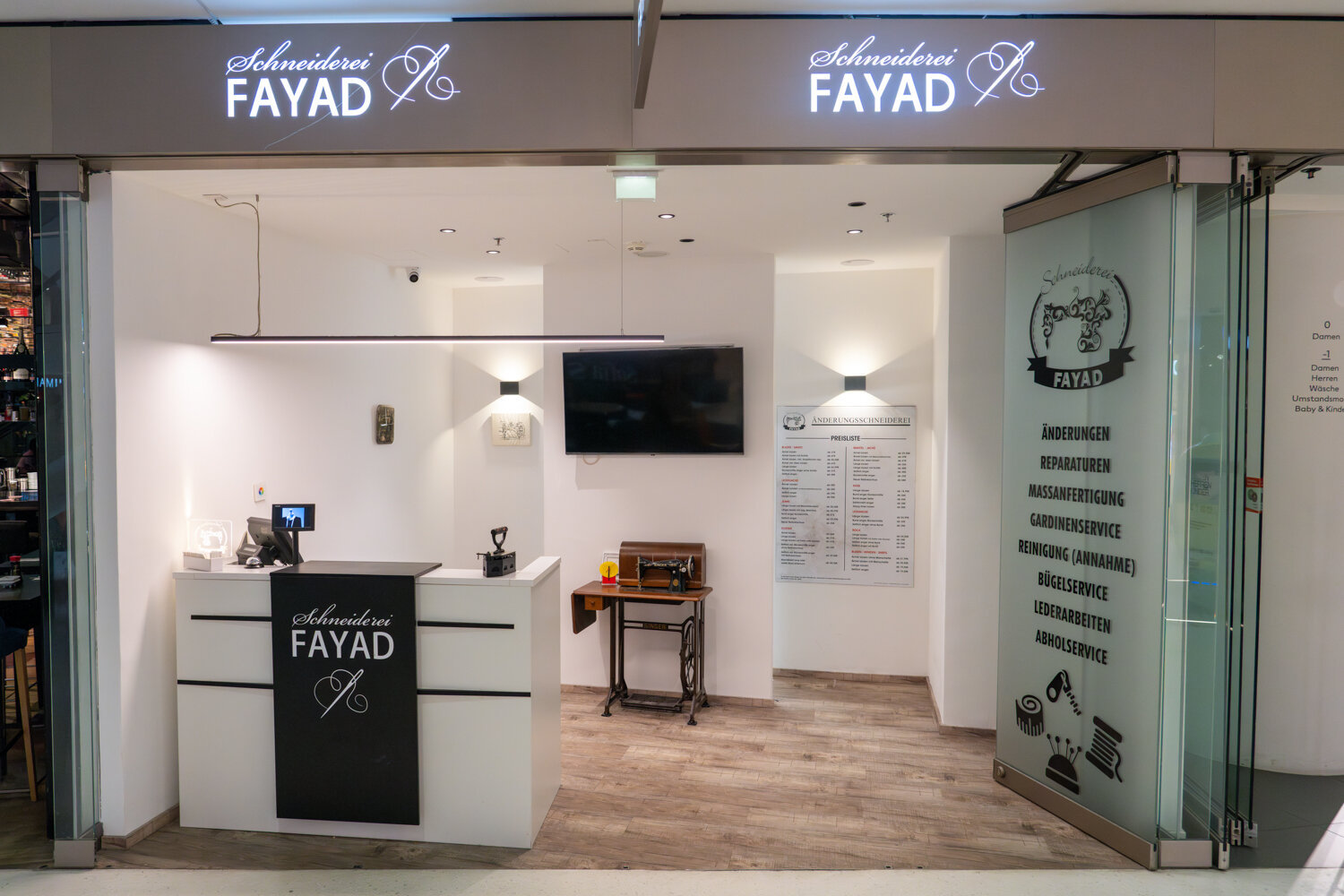 Eingangsbereich der Schneiderei Fayad mit Rezeptionstheke, historischer Nähmaschine und Preisliste an der Wand.