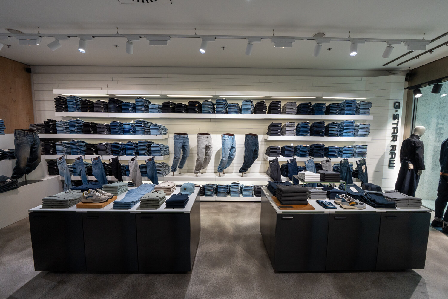 Frontalansicht der großen G-Star RAW Jeans-Wand mit aufgereihten Denim-Hosen, Präsentations-Displays und zwei schwarzen Tischinseln voller Kleidung.