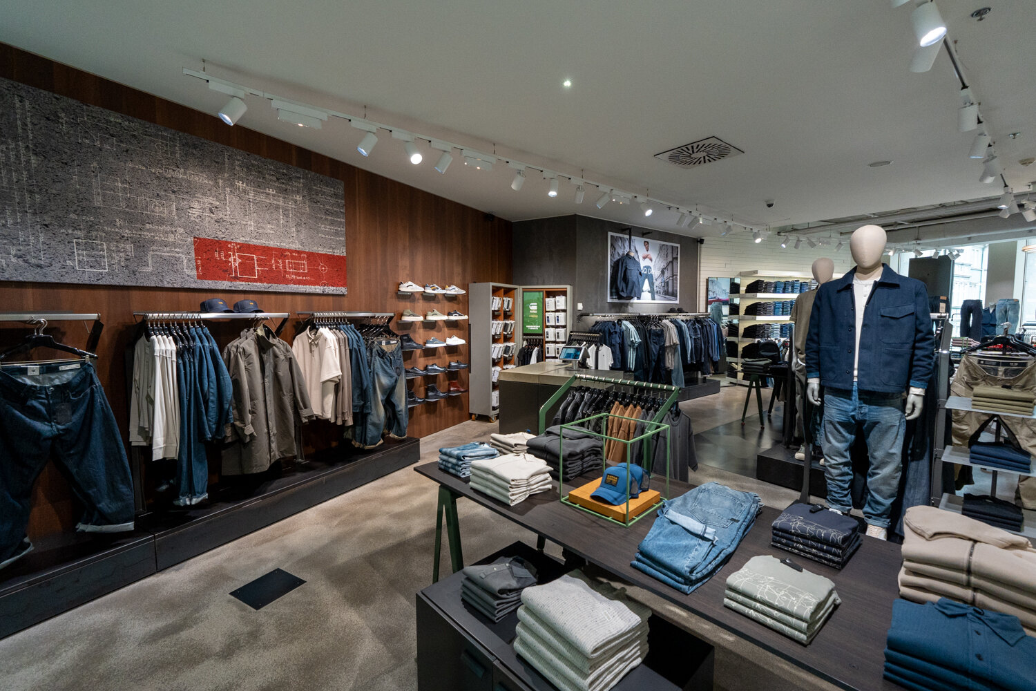 Verkaufsraum von G-Star RAW mit eingekleideter Schaufensterpuppe, Kleiderständern an einer Holz-Wandverkleidung und Tischen voller gefalteter Kleidung.