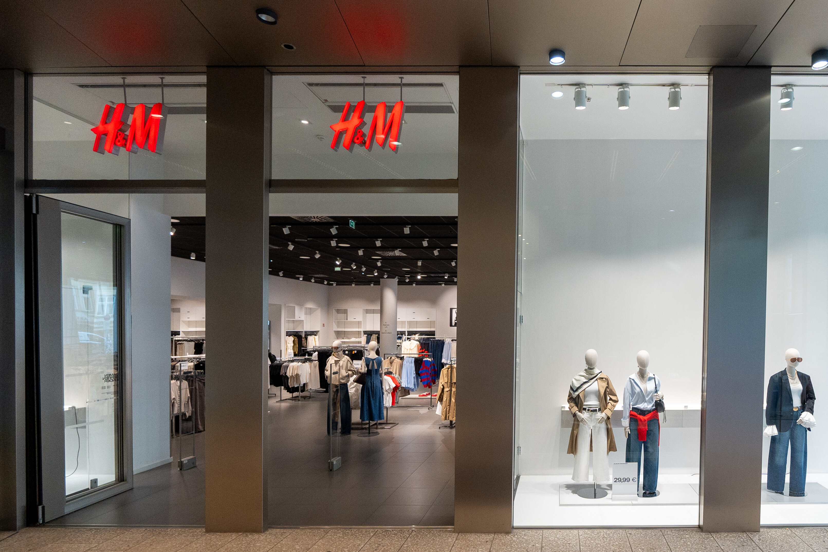 Eingangsbereich des H&M-Geschäfts im Kaufhaus Tyrol mit leuchtendem roten Markenlogo und drei Schaufensterpuppen im Schaufenster.
