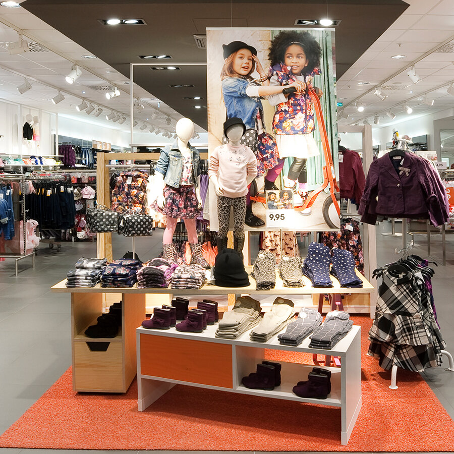 Farbenfroher Präsentationstisch in der Kinderabteilung mit Schaufensterpuppen, ordentlich gefalteter Kleidung und kleinen Schuhen auf einem orangen Teppich.