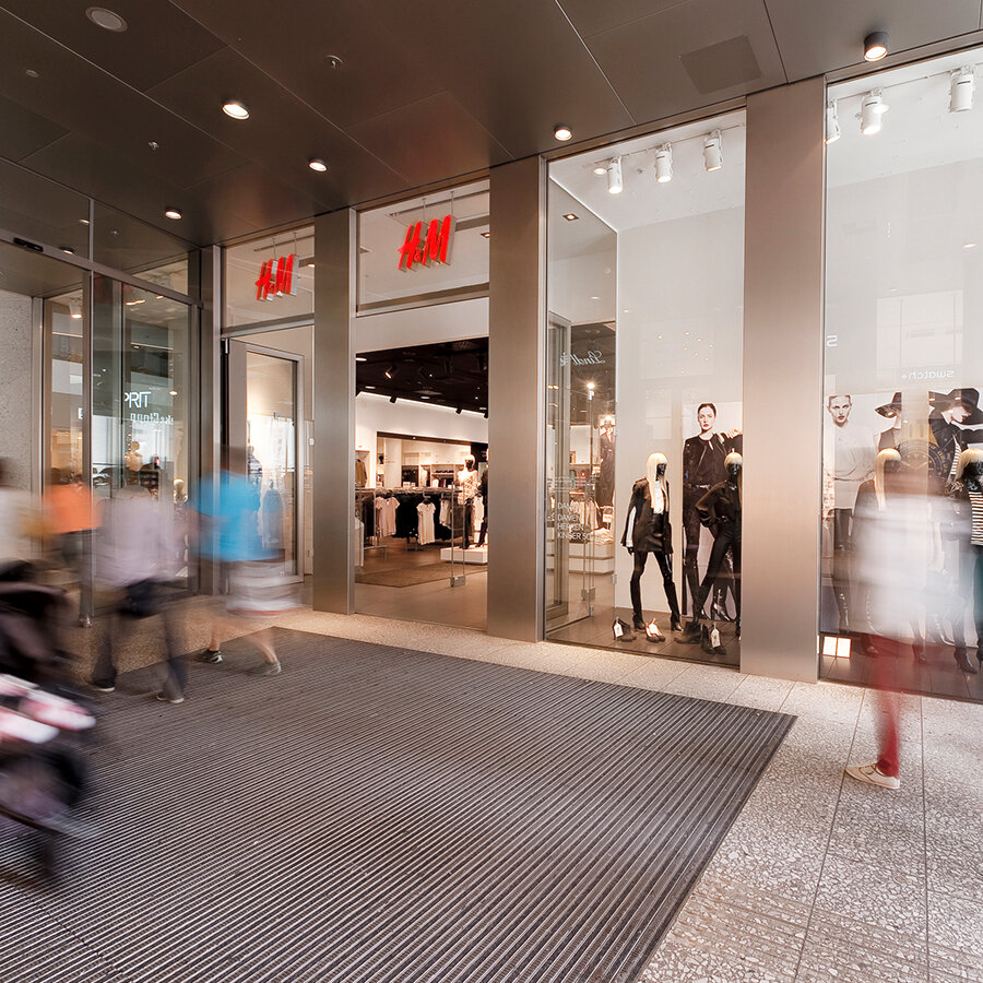 Außenansicht der H&M-Filiale im Einkaufszentrum mit großen Glasfronten, beleuchteten Logos und vorbeigehenden Passanten im Bewegungsunschärfe-Effekt.