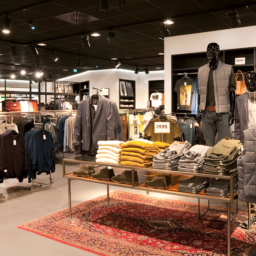 Gut sortierte Herrenabteilung bei H&M mit eleganten Sakkos, gefalteten Strickpullovern auf einem Glastisch und einem dekorativen Teppich.