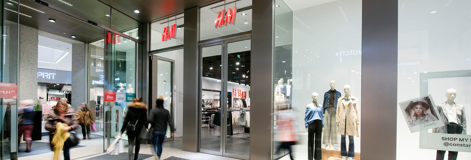 Weitläufiges Breitbild-Panorama des H&M-Filialzugangs im Kaufhaus Tyrol mit gläserner Front und aktuellen Modetrends im Schaufenster.