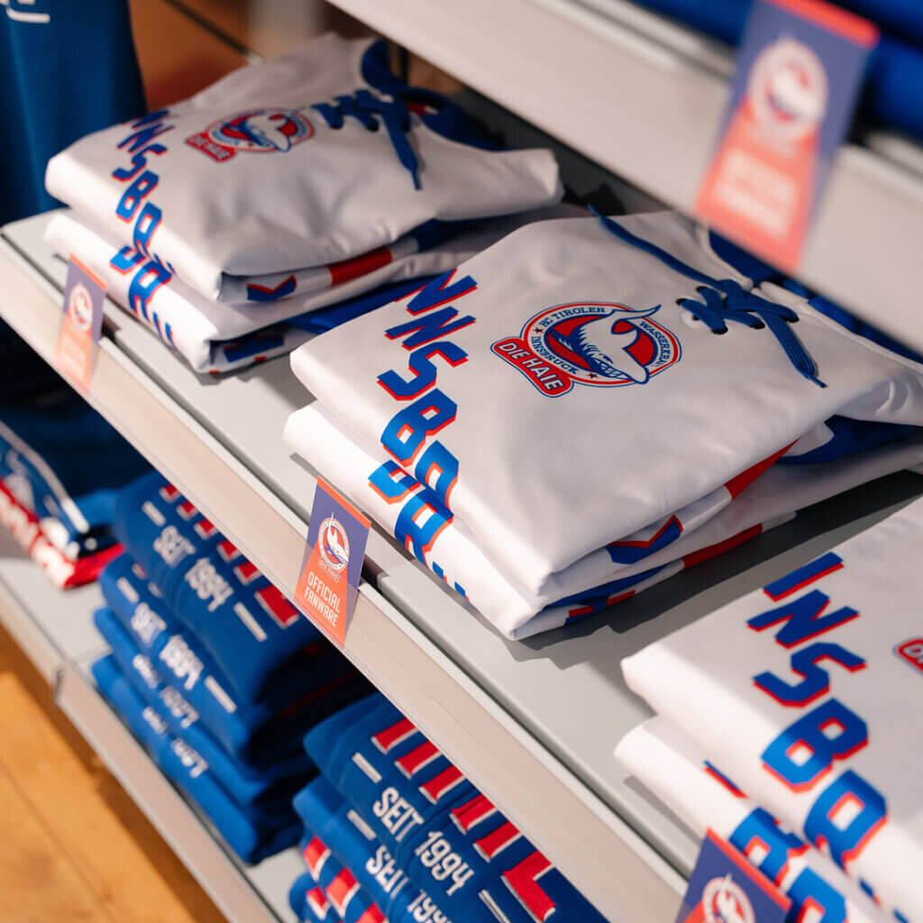 Ordentlich gestapelte, weiße und blaue Fantrikots mit dem Logo der Innsbrucker Haie im Regal des Fan-Shops.
