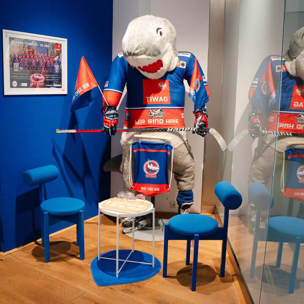 Lebensgroße Hai-Maskottchen-Figur in voller Eishockey-Montur mit Schläger inmitten einer blauen Sitzecke für Fans.