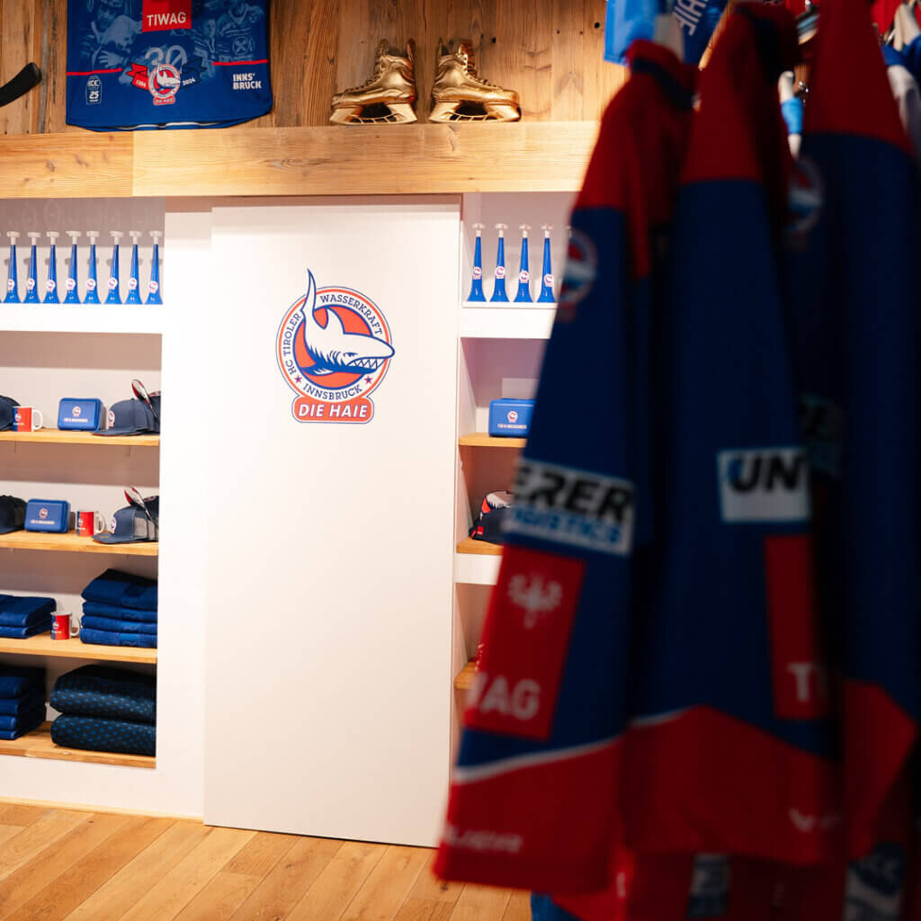 Einblick in den Fanshop mit Kappen, Tassen und hängenden Eishockey-Trikots vor einer gemütlichen Holzwand mit goldenen Schlittschuhen.