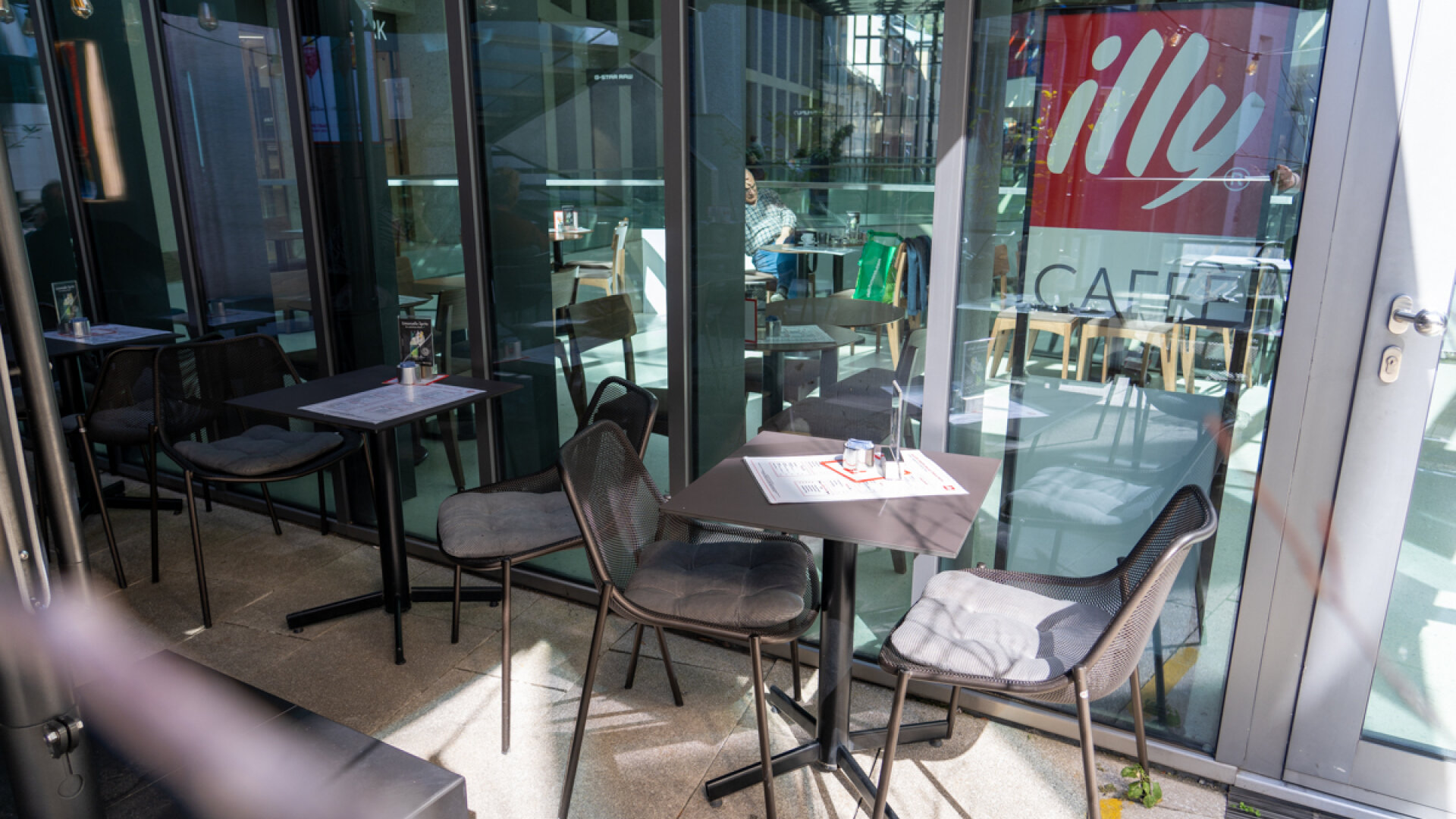 Gemütlicher Sitzbereich vor den großen Fensterfronten des illy Cafés mit leuchtendem Markenlogo.