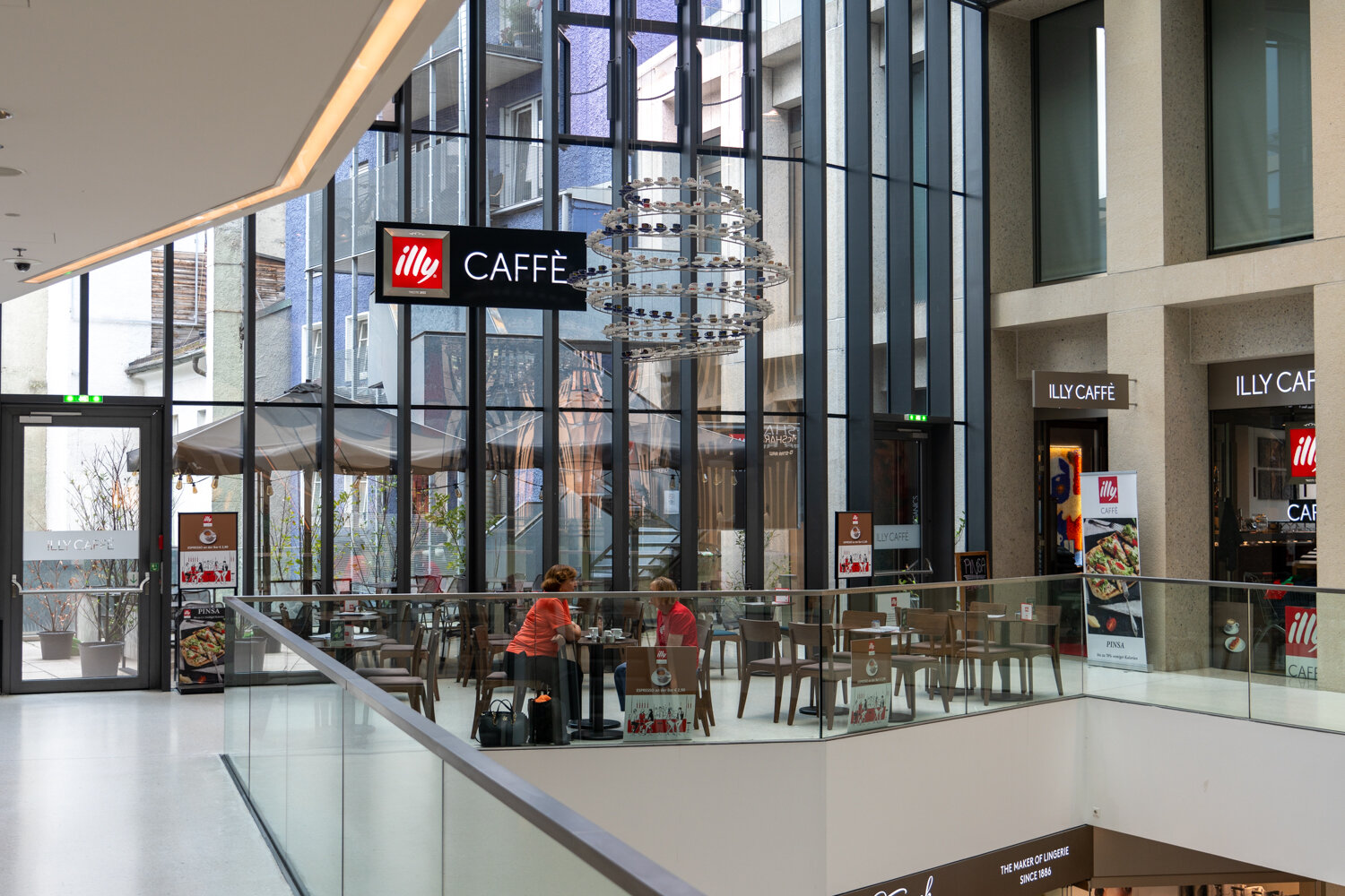 Sitzbereich von Illy Caffè auf der Galerie im Einkaufszentrum mit Gästen, Tischen und gläserner Fassade.