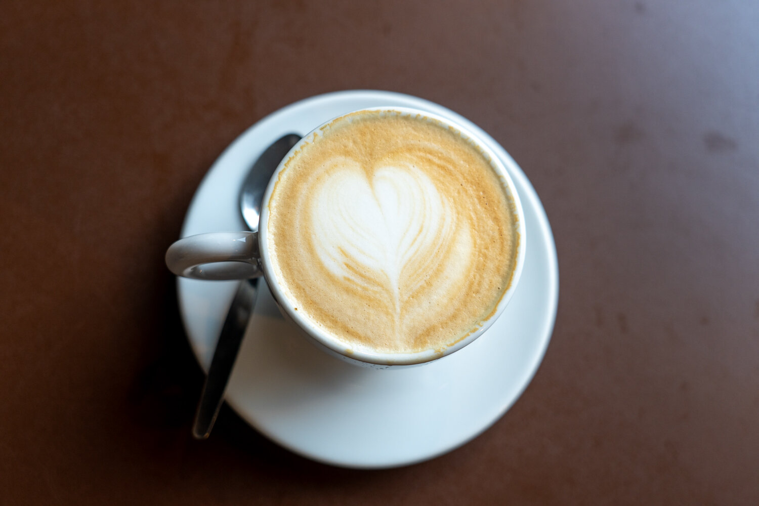 Tasse Cappuccino mit Schaumherz von oben fotografiert auf Untertasse und braunem Tisch.