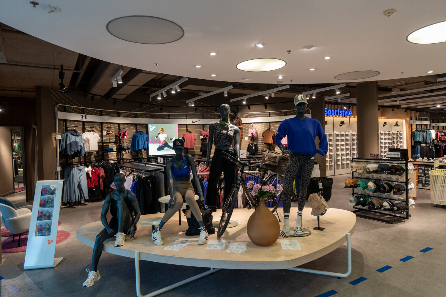 Ausstellungsplattform bei Intersport mit mehreren dunkelgrauen Schaufensterpuppen in Sportkleidung, einem Fahrrad Mützenständer und Wandregalen mit Schuhen.
