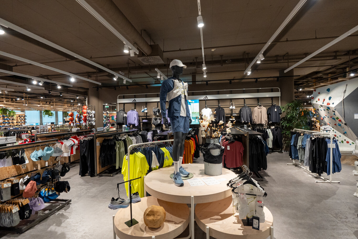 Verkaufsfläche bei Intersport mit Schaufensterpuppe im Lauf-Outfit auf hölzernem Podest, Sportbekleidung an Ständern und einer Kletterwand im Hintergrund.