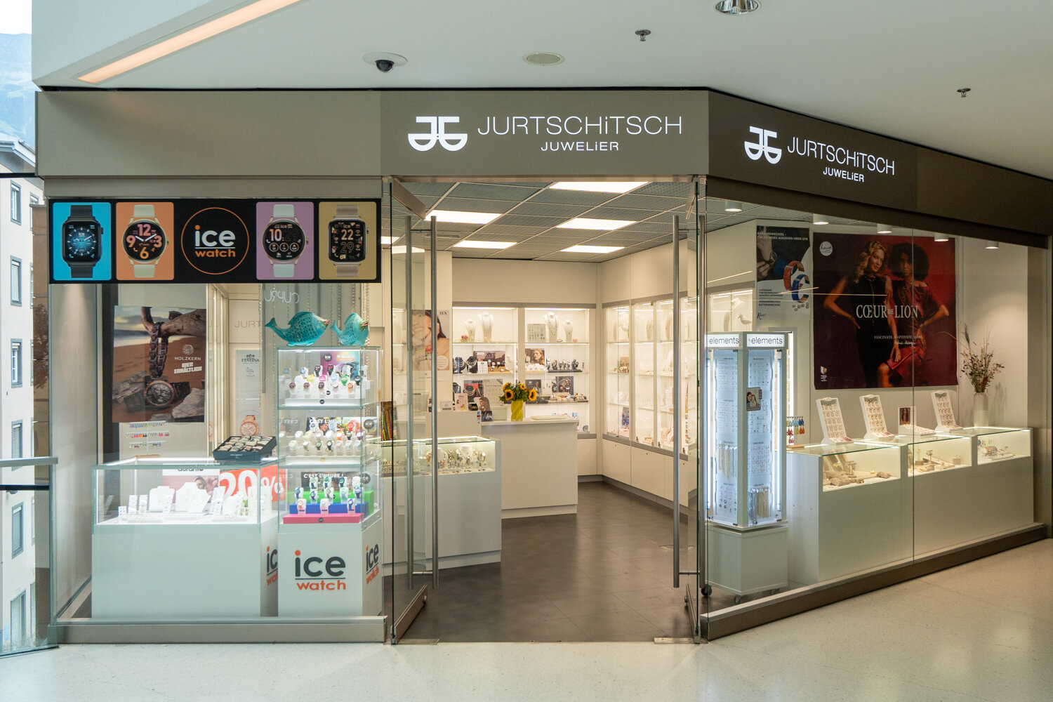 Frontalansicht der Filiale Jurtschnitsch Juwelier mit beleuchtetem Logo, Glasvitrinen für Ice-Watch Uhren, Schmuck-Displays und Einblick in den Verkaufsraum.