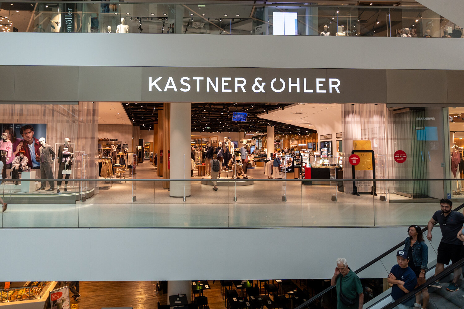 Eingang zur Kastner & Öhler Filiale mit leuchtendem Schriftzug Glasgeländer im Vordergrund und Einblick in die Mode- und Kosmetikabteilung.