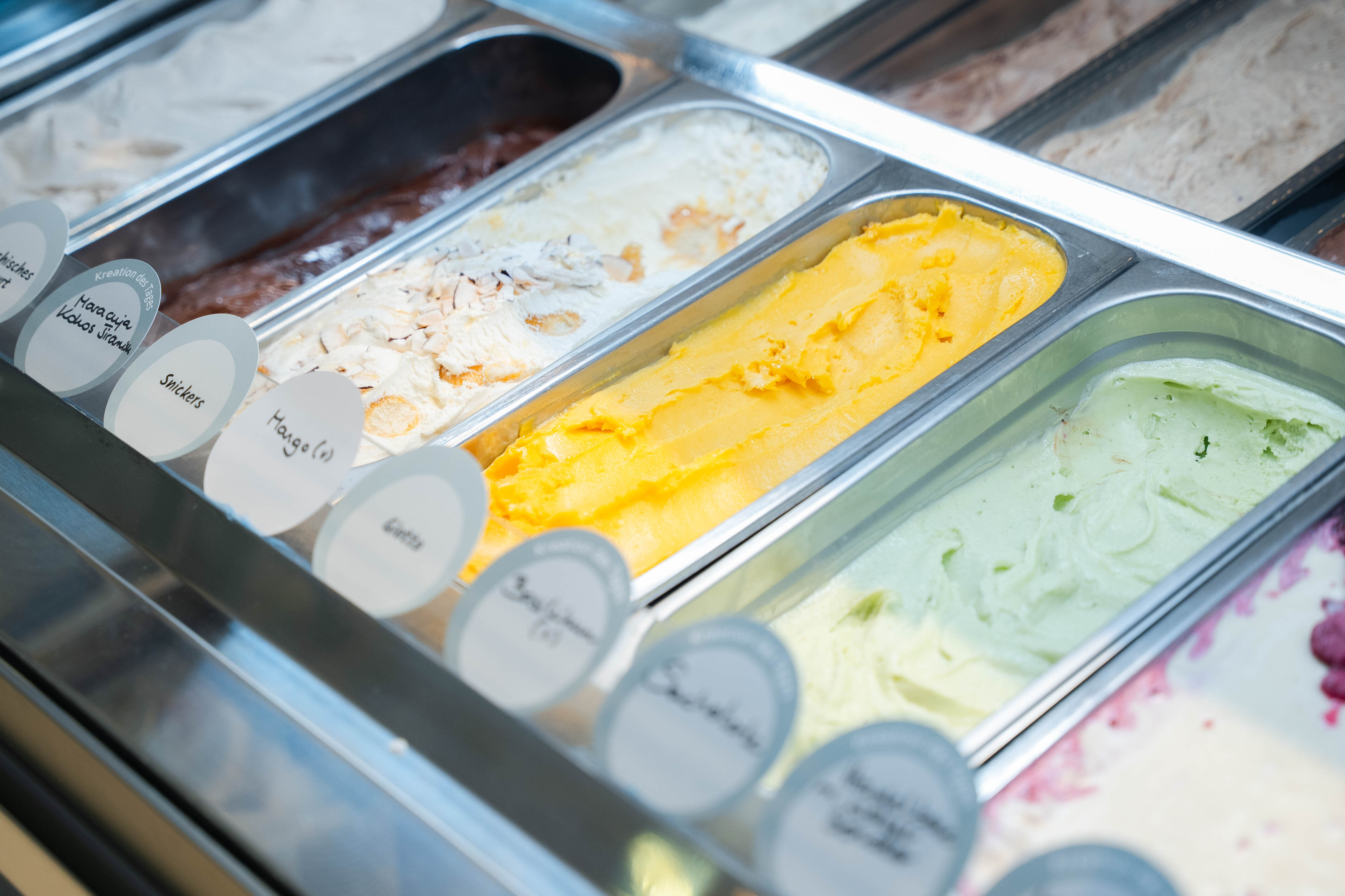 Nahaufnahme einer Eisvitrine mit verschiedenen Eissorten wie Mango, Giotto und Pistazie in Edelstahlbehältern.