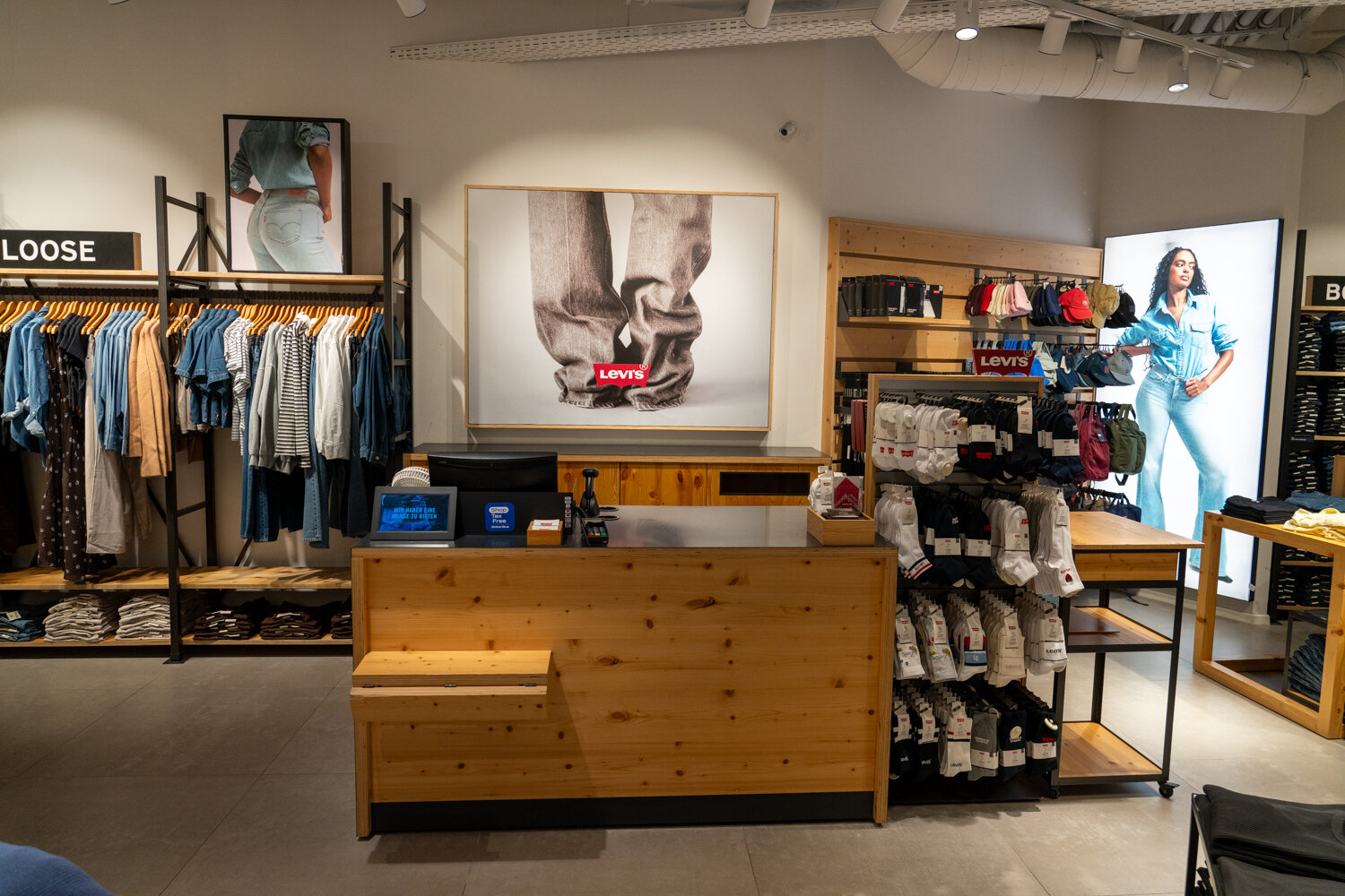 Holztresen im Kassenbereich des Levi’s Stores mit Werbepostern, Sockenständer und Hängebekleidung im Hintergrund.