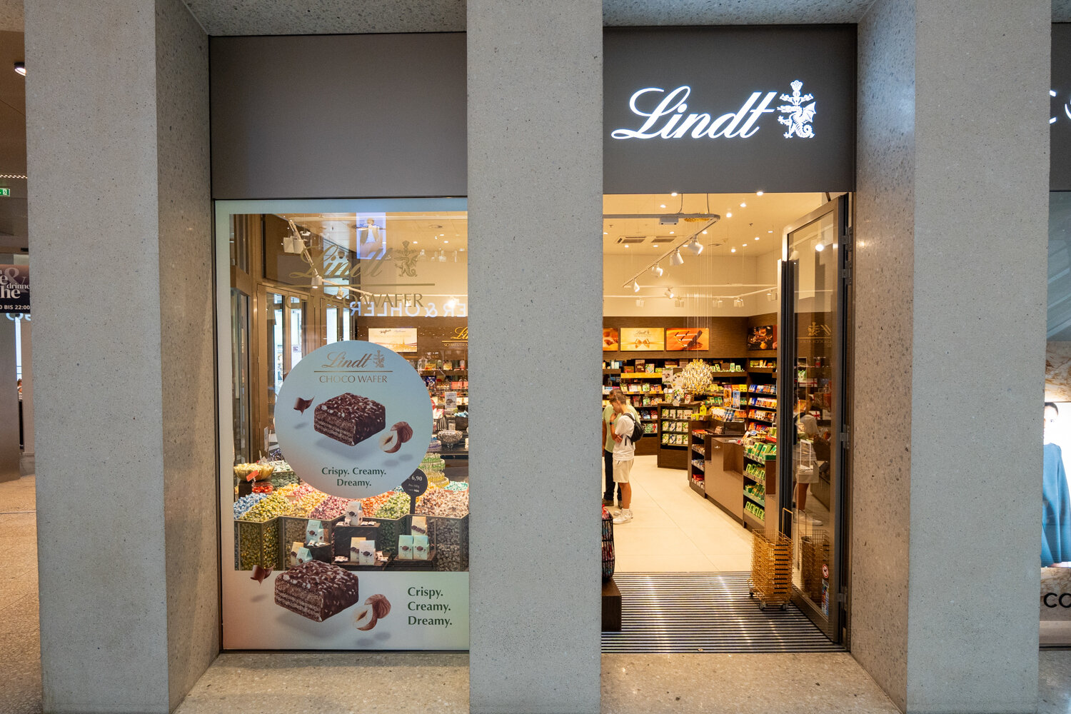 Frontalansicht der Lindt Filiale mit beleuchtetem Logo über dem Eingang, Schaufenster-Plakat für Choco Wafer und Einblick in das Schokoladengeschäft.