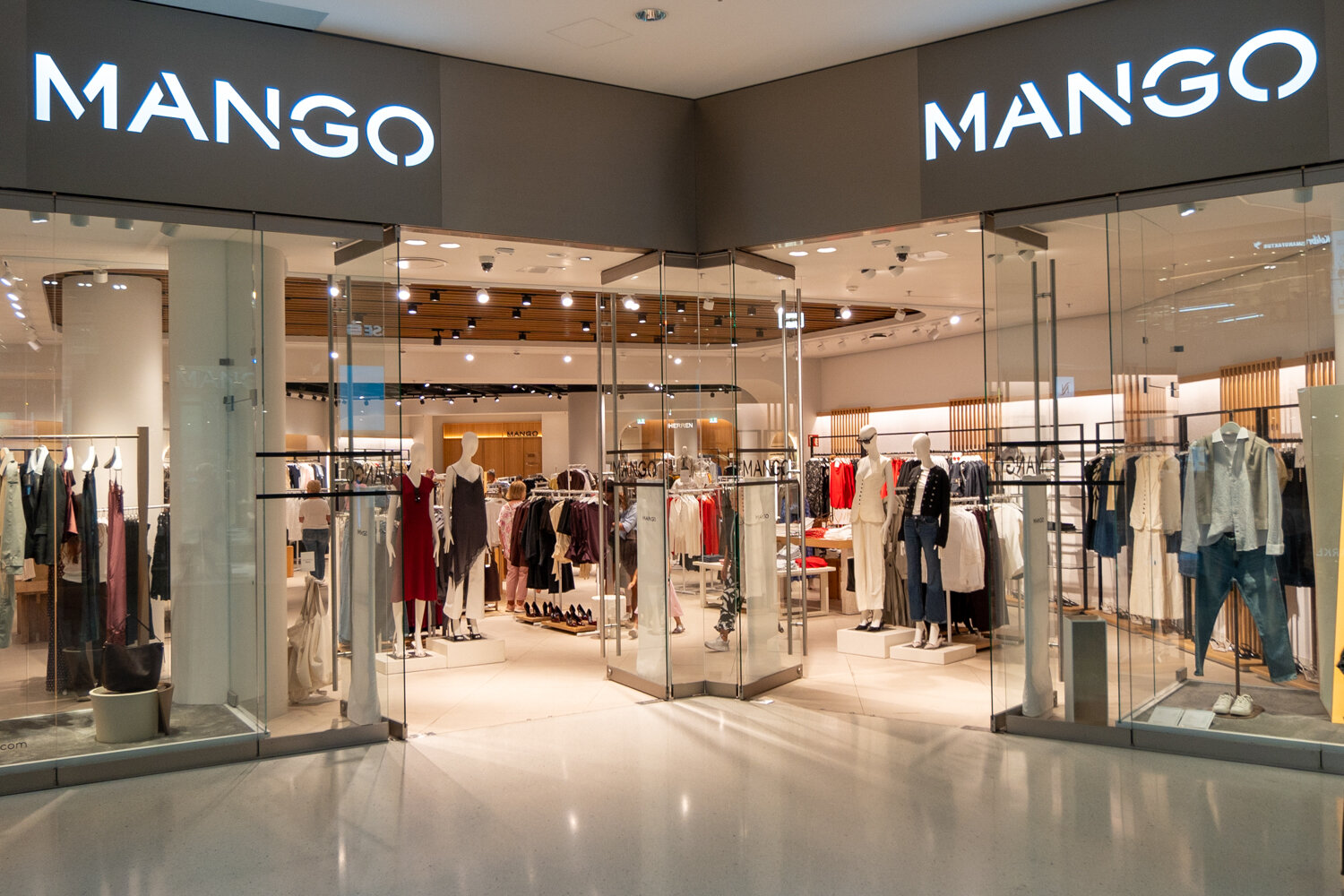 Große Frontansicht der Mango Filiale mit zwei beleuchteten Logos, Glaseingangstüren, Schaufensterpuppen in Damenbekleidung und Einblick in den Verkaufsraum.