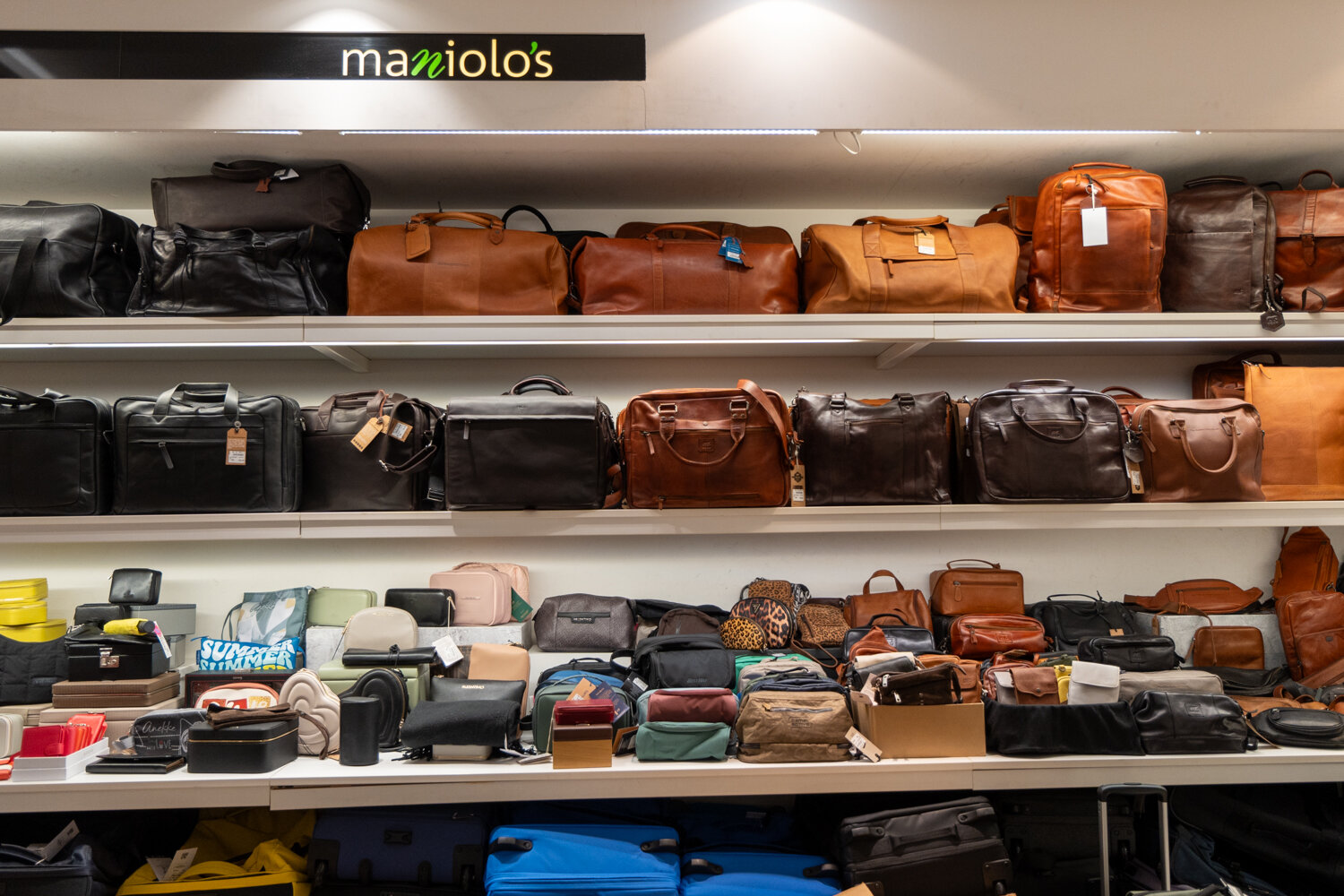 Beleuchtetes Wandregal unter dem Logo Maniolo's mit Laptoptaschen, Aktentaschen, Reisetaschen aus Leder, Kulturbeuteln und Koffern im unteren Bereich.