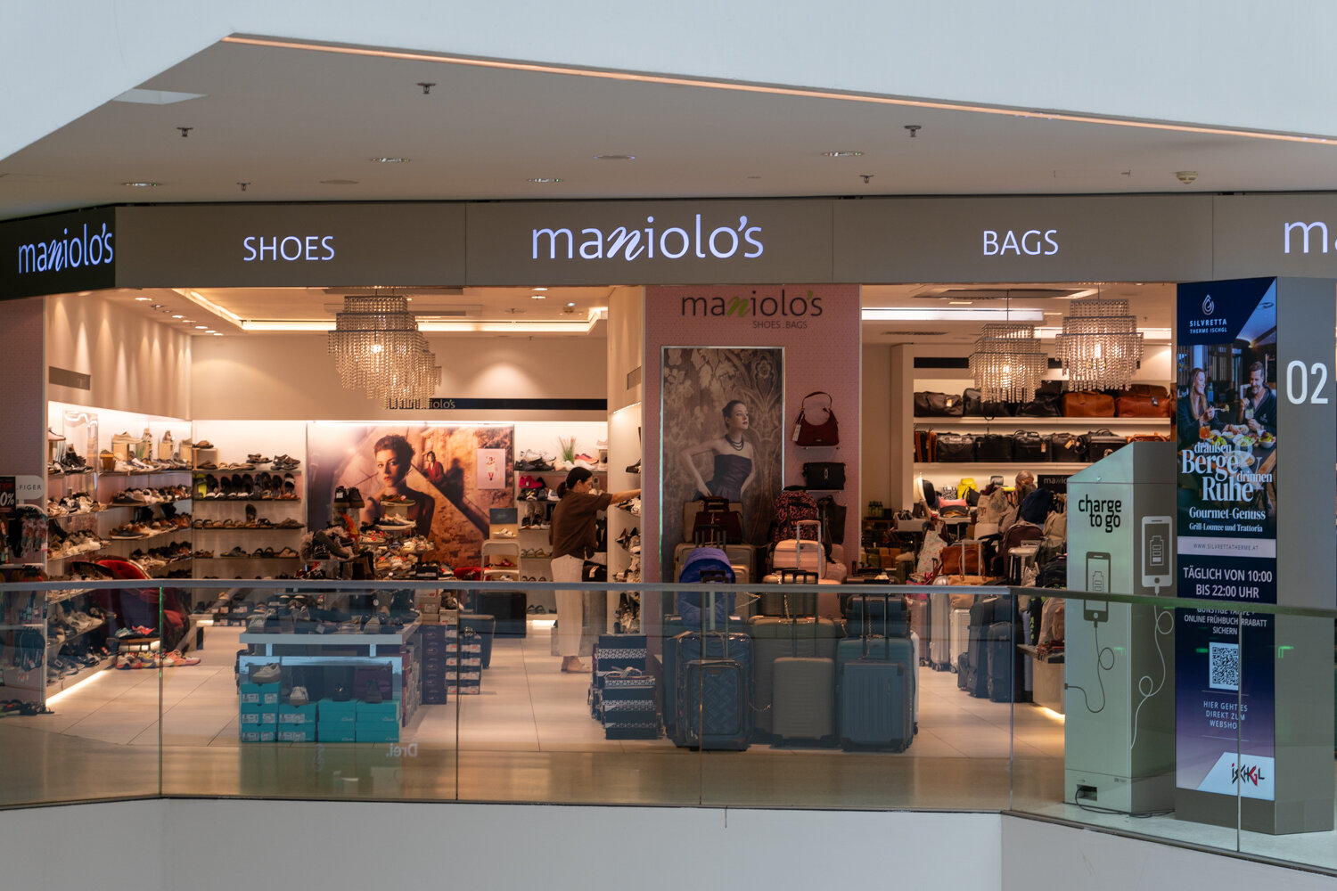 Frontansicht des Geschäfts Maniolos Shoes & Bags im Einkaufszentrum mit Koffer-Präsentation, Schuhregalen und Beleuchtung.