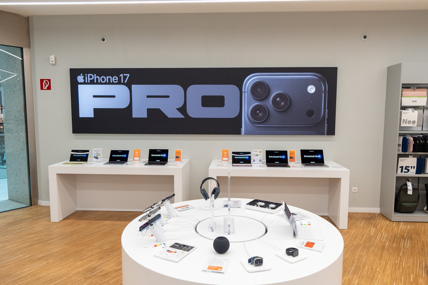 Runder Präsentationstisch bei McSHARK mit Kopfhörern und Apple Watch Zubehör vor Tischen mit MacBooks und einem großen iPhone 17 Pro Wandtransparent.