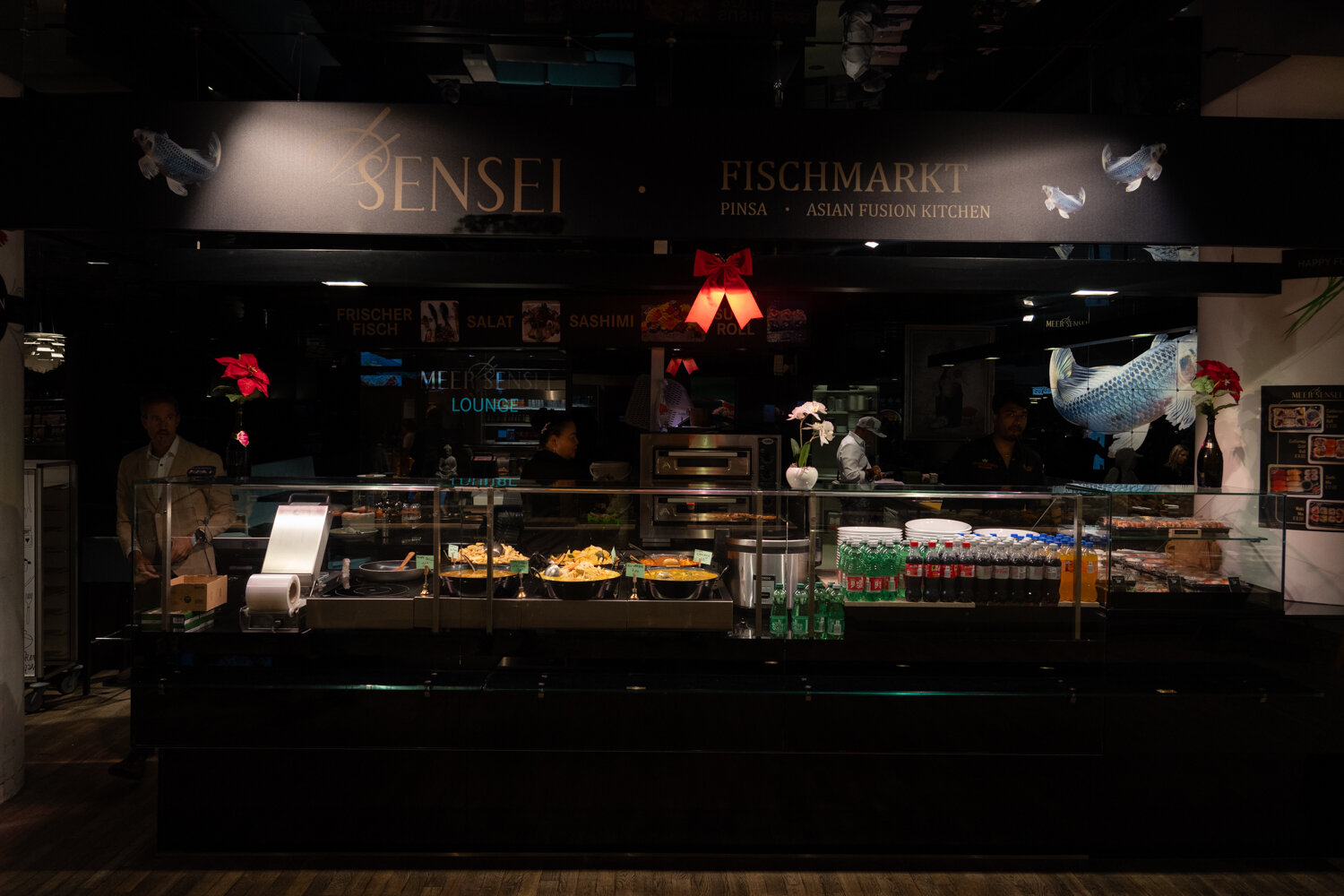 Frontansicht der Restauranttheke von Meer Sensei Fischmarkt mit warmen asiatischen Gerichten, Getränken und beleuchtetem Logo.