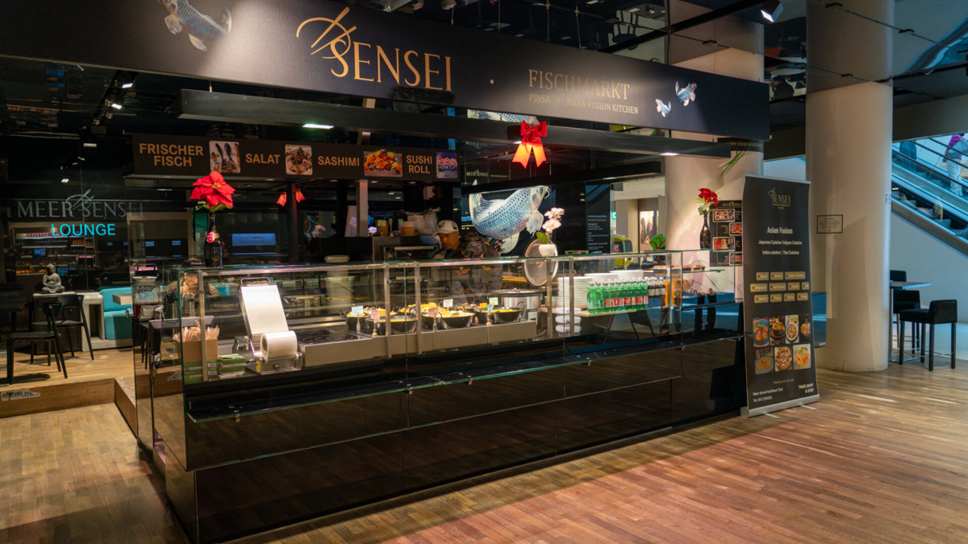 Frontansicht der Restauranttheke von Meer Sensei Fischmarkt mit warmen asiatischen Gerichten, Getränken und beleuchtetem Logo.