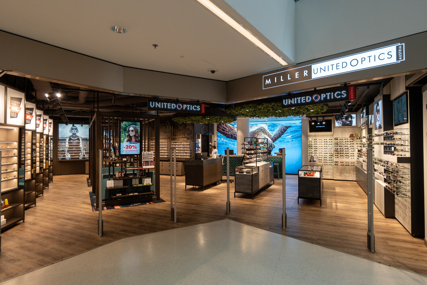 Frontansicht eines modernen Miller United Optics Optikergeschäfts im Einkaufszentrum mit Holzoptik-Boden und beleuchteten Brillendisplays.