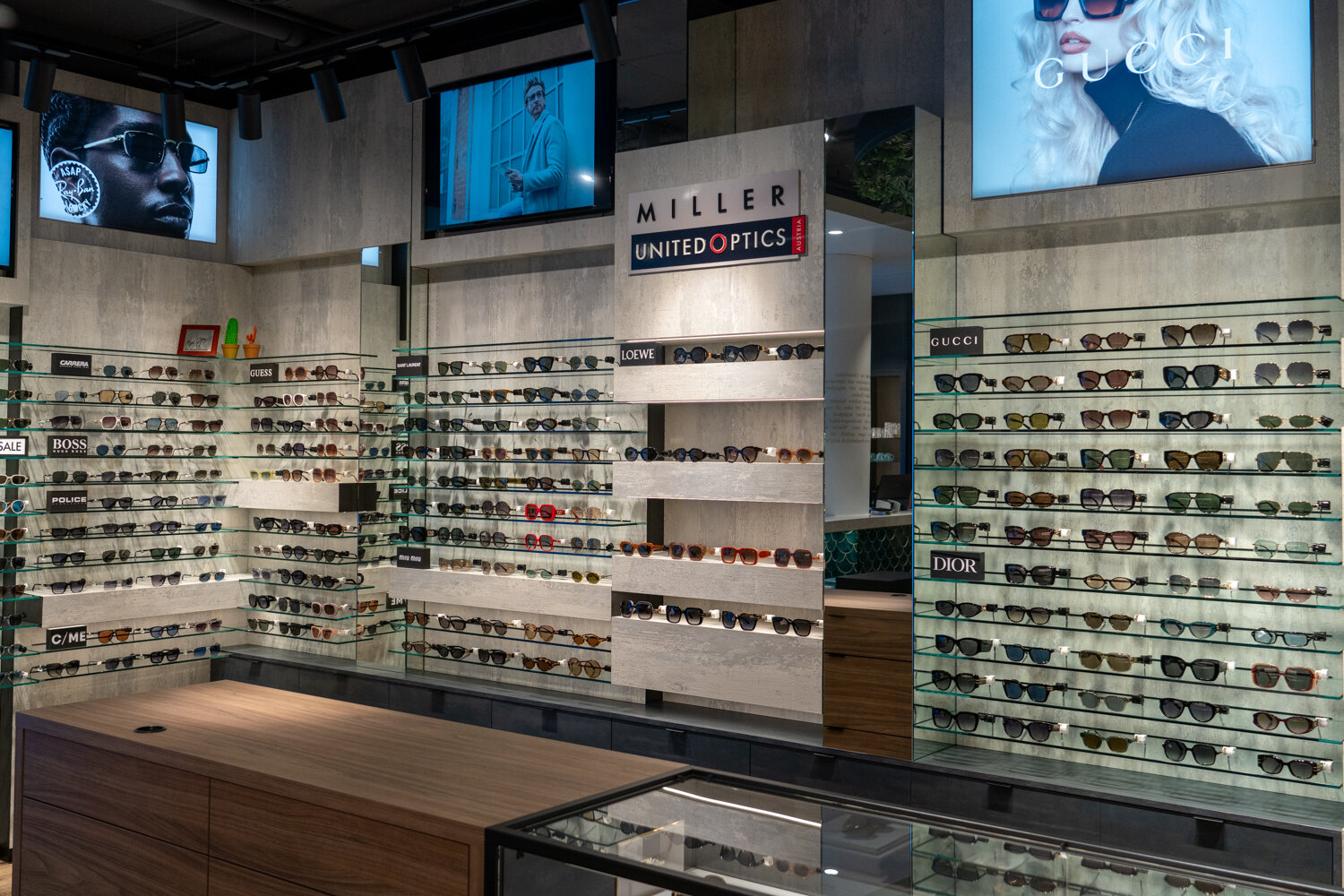 Designer-Sonnenbrillen-Wand bei Miller United Optics mit Glasregalen, Verkaufstresen und Werbedisplays von Gucci, Dior und Loewe.