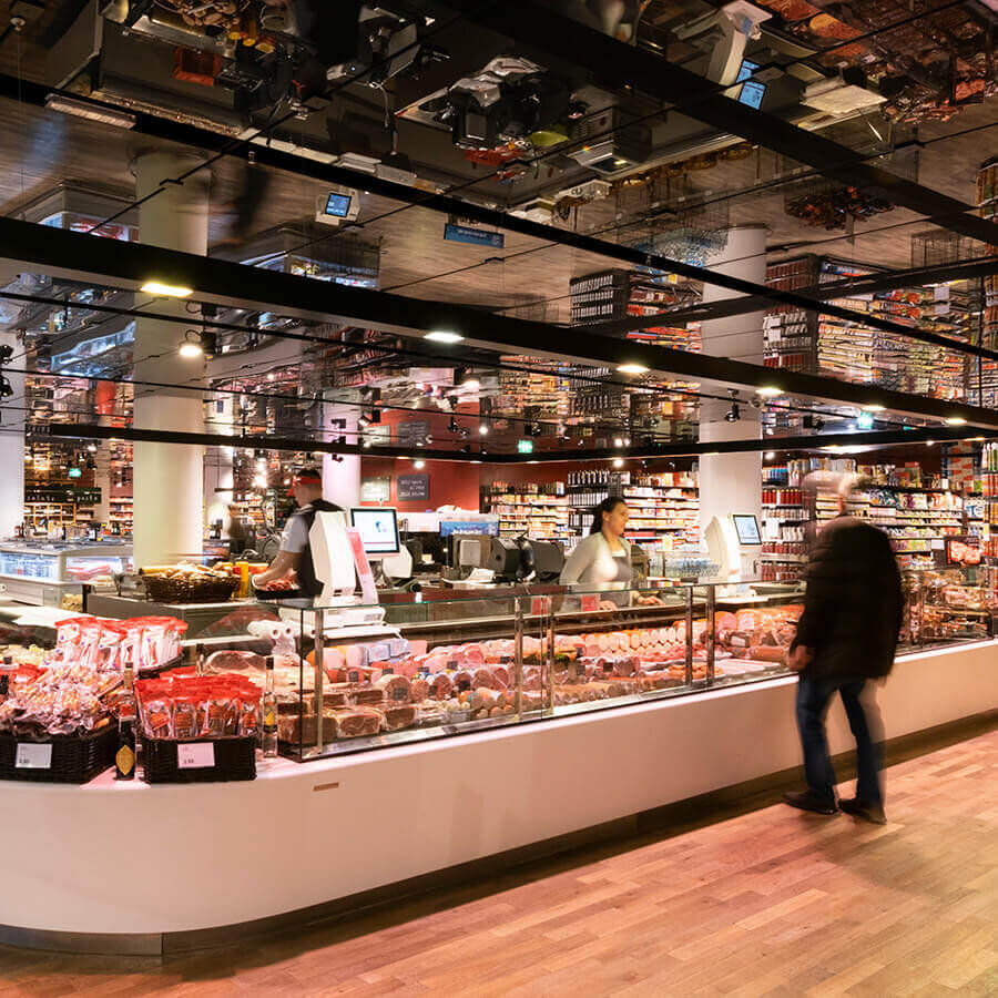 Eine lange, hell erleuchtete Fleisch- und Wursttheke in einer MPREIS-Filiale. Zwei Mitarbeiter bedienen hinter der Glasvitrine, während ein Kunde an der Theke vorbeigeht. Das moderne Ladendesign kombiniert dunkle Metallelemente, warmes Holz und eine markante Spiegeldecke.