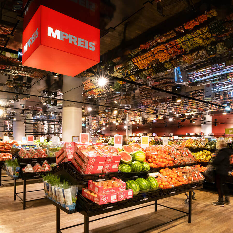 Die bunte Obst- und Gemüseabteilung eines MPREIS-Marktes mit Kisten voller Wassermelonen, Orangen, Äpfel und frischen Kräutern. Über den Auslagen hängt ein großer, leuchtend roter MPREIS-Logowürfel. Der gesamte Frischebereich spiegelt sich effektvoll in der dunklen Hochglanzdecke.