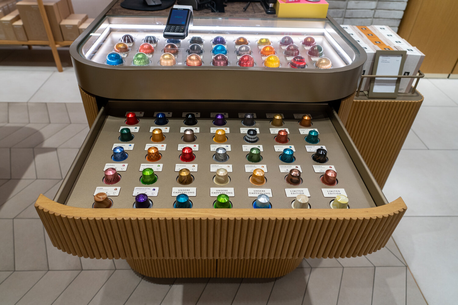 Ausgezogene Holzschublade an einer Präsentationstheke mit sortierten, bunten Nespresso-Kaffeekapseln.