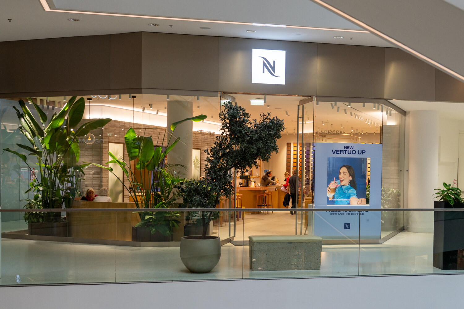 Glasfassade der Nespresso Boutique im Kaufhaus Tyrol mit leuchtendem N-Logo, großen Zimmerpflanzen und Werbedisplay.