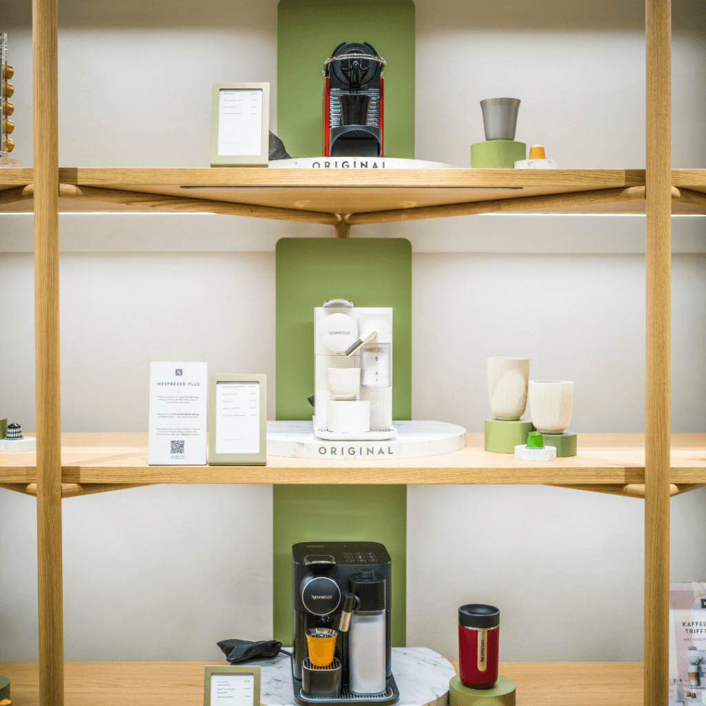 Elegantes Holzregal mit verschiedenen Nespresso Original Kaffeemaschinen und farblich passenden Kaffeetassen auf grün-weißen Design-Podesten.