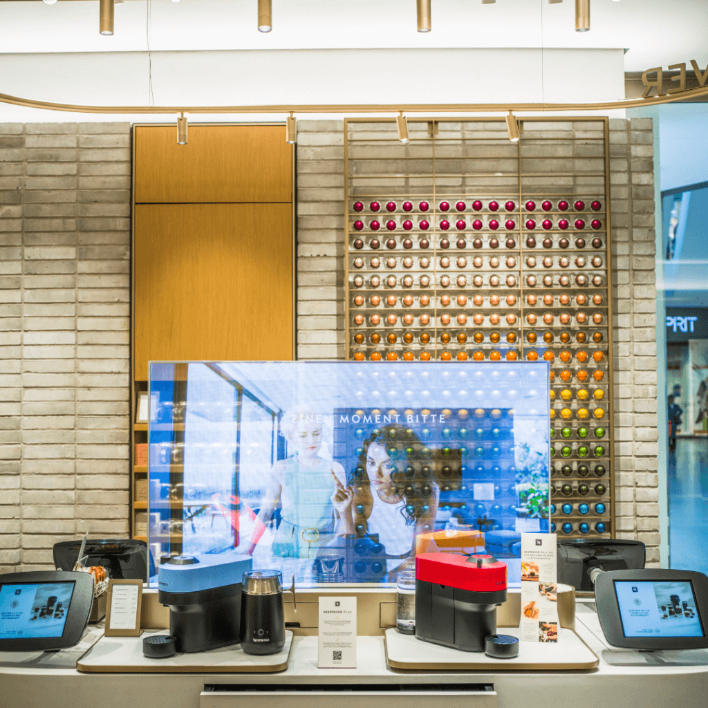 Moderner Verkostungstresen in der Nespresso Boutique mit bunten Kaffeemaschinen, Milchaufschäumer und einer riesigen Kaffeekapsel-Wand im Hintergrund.