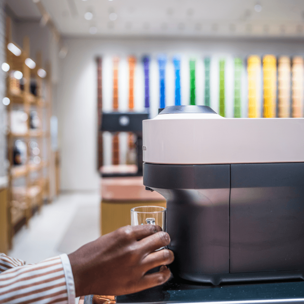 Eine Hand hält eine edle Glastasse unter den Kaffeeauslauf einer modernen, weißen Nespresso Vertuo Kaffeemaschine während des Brühvorgangs.