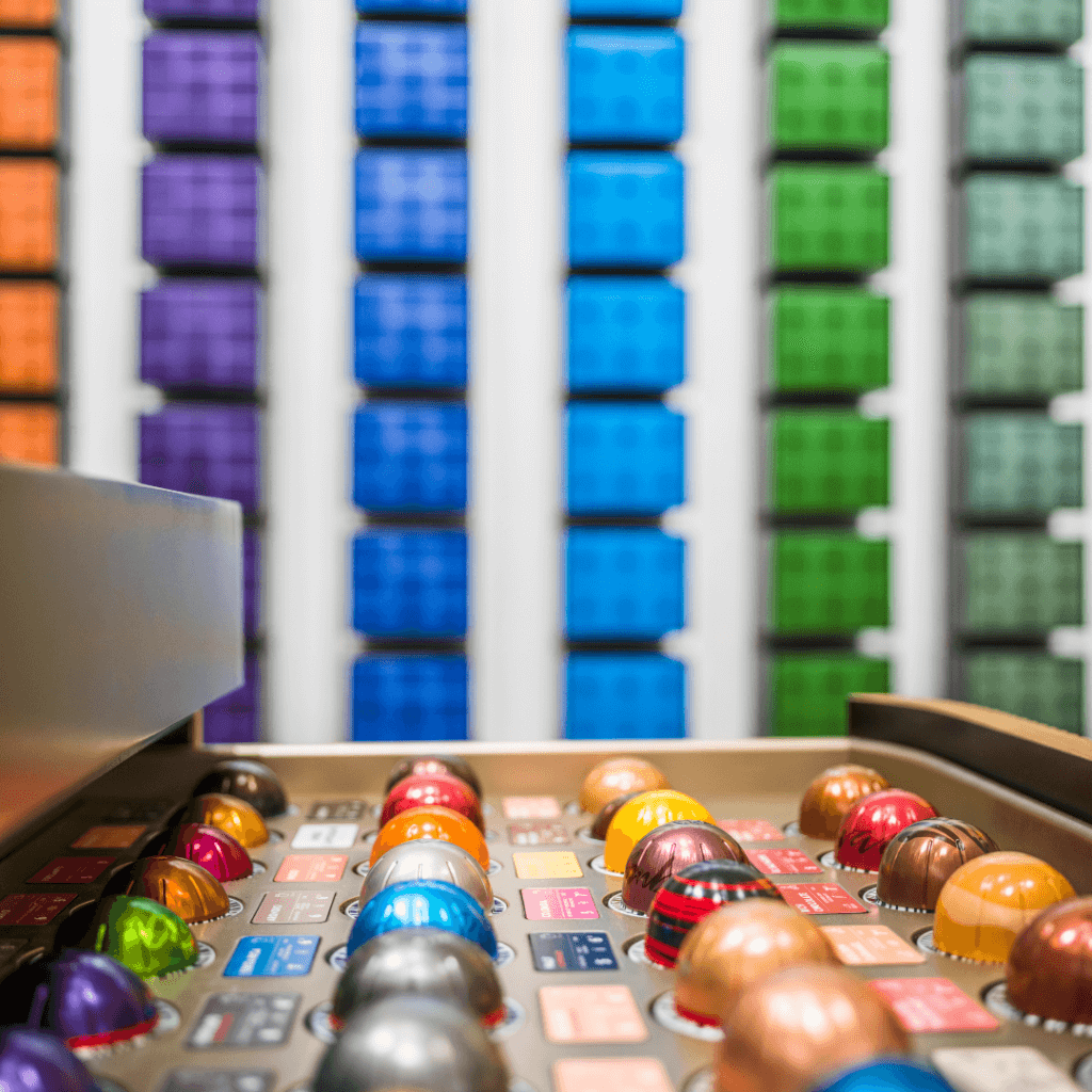 Nahaufnahme einer bunten Auswahl an Nespresso Kaffeekapseln in einer Präsentationsschale vor einer Wand aus farblich sortierten Kapsel-Verpackungen.