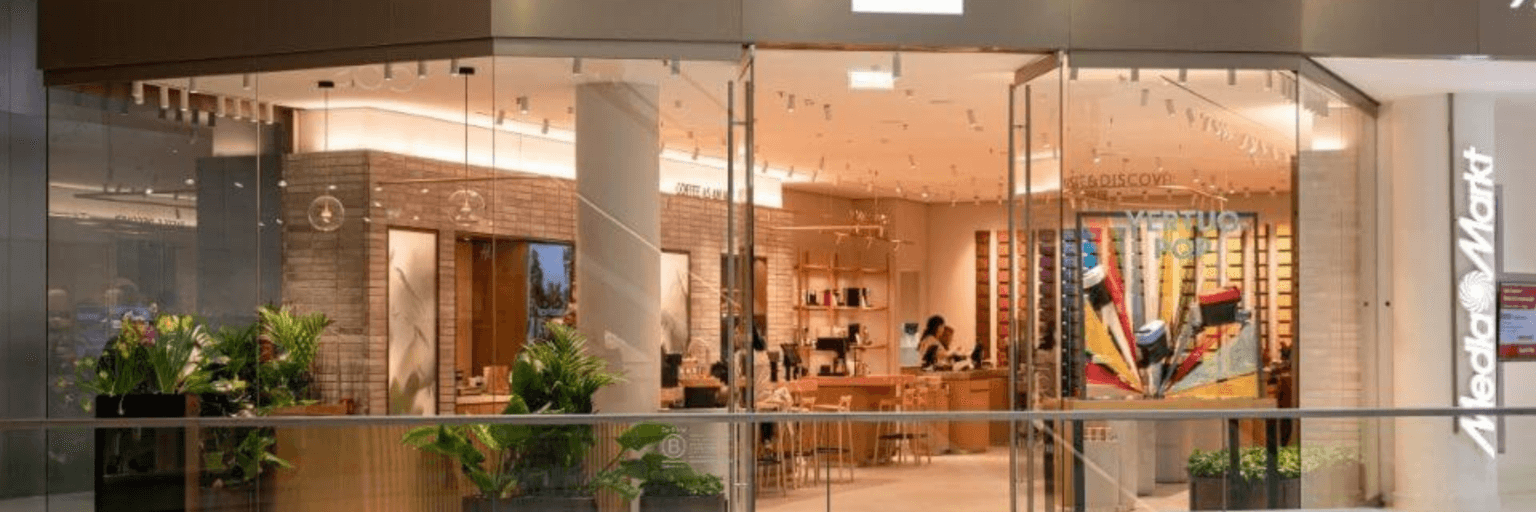 Weitläufiges Breitbild-Panorama der modernen Nespresso Boutique im Kaufhaus Tyrol mit stilvoller Holzeinrichtung, großen Glasfronten und grünen Zimmerpflanzen.