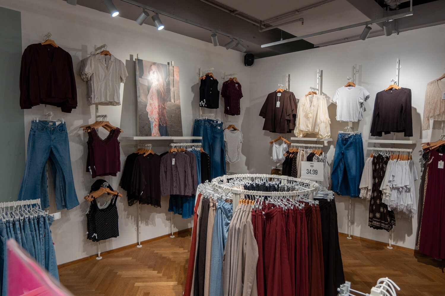Verkaufsraum eines ONLY Bekleidungsgeschäfts mit hängender Damenmode, Jeans, Oberteilen und Rundleiderständern auf Fischgrätparkett.