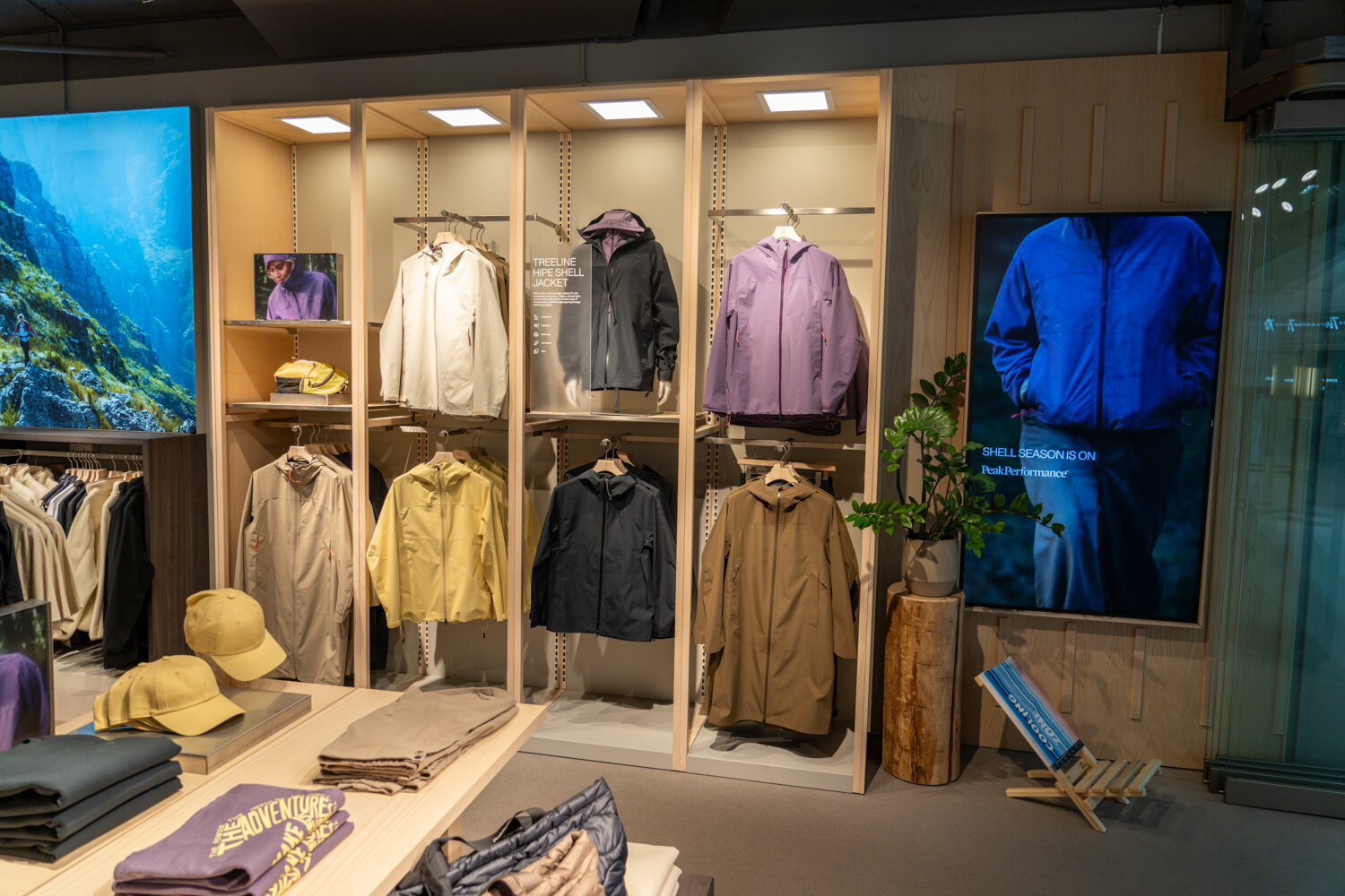 Wandregal aus hellem Holz bei Peak Performance mit wetterfesten Jacken und Mänteln unter der Beschilderung Treeline Hipe Shell Jacket.